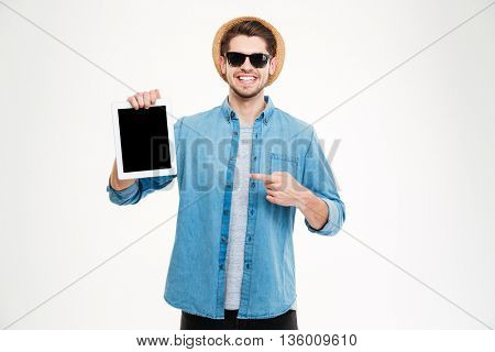 Cheerful young man in sunglasses and hat holding and pointing on blank screen tablet over white background