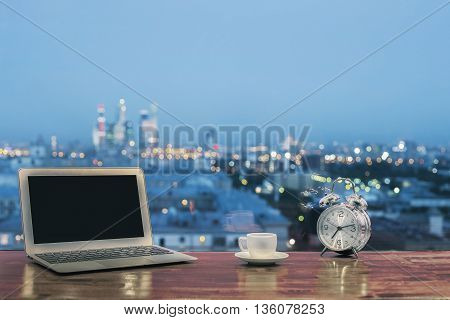 Front view of creative designer desktop with blank laptop computer silver alarm clock and coffee cup on blurry night city background. Mock up