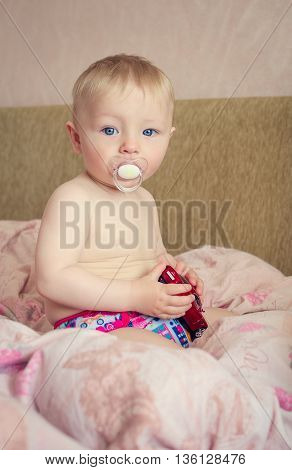 Adorable naked blond baby boy with blue eyes sitting and playing with a toy car in the parents bed. Cute toddler in pink diaper with pacifier. Beautiful baby is sitting on a bed.