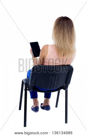 back view woman sitting on chair and looks at the screen of tablet. Rear view people collection.  Blonde in blue trousers sitting on the office chair and reading with a tablet computer.