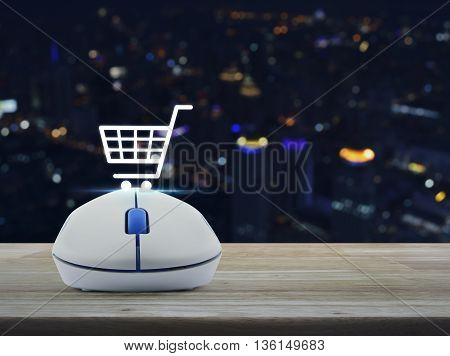 Wireless computer mouse with shopping online icon on wooden table in front of blurred light city tower Shopping online concept