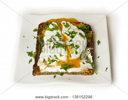 A poached egg freshly cooked and cut on a piece of rye toast with fresh herbs on top on a white plate on white background
