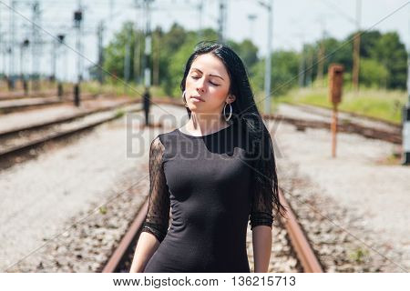 Half length of a young pretty girl in black dress and nylons walking down rail tracks, cargo wagons in background, eyes closed