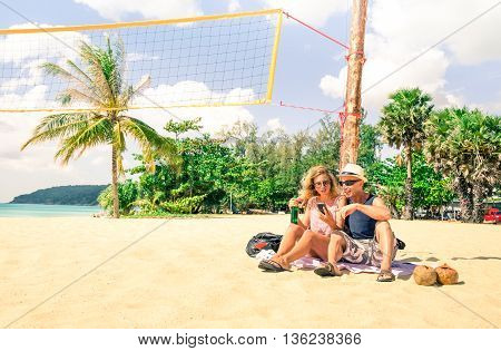 Tourist couple using mobile phone on the beach at Phuket Island - Travel friends looking holiday pictures sitting on the white sand - Concept of relax and technology in a tropical destination tour