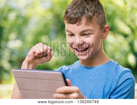Outdoor portrait of happy teen boy 12-14 year old with tablet pc watching video in summer park