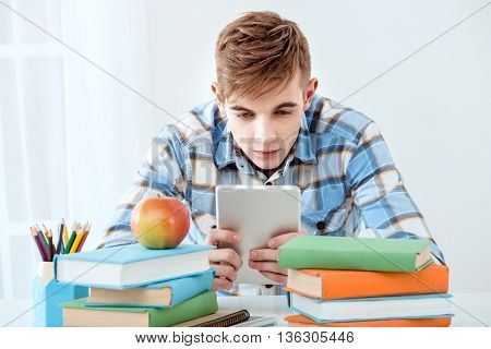 Modern problem: youth and computers. Young student using tablet computer while studying. Nice white interior with table full of books