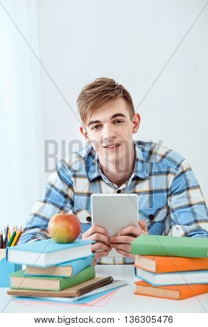 Modern problem: youth and computers. Young student using tablet computer while studying and looking at camera. Nice white interior with table full of books