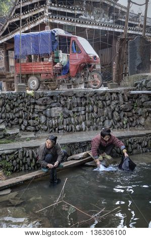 Zhaoxing Dong Village Guizhou Province China - April 8 2010: Two Chinese women rinse laundry in water countryside river near the cobblestone the seafront and parked motor tricycle.