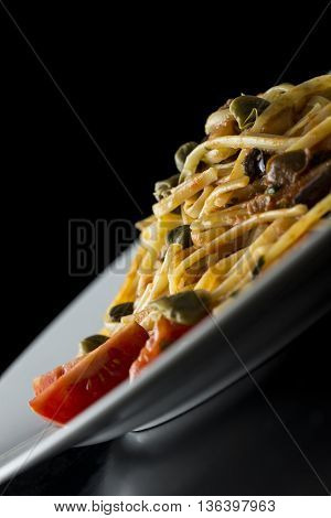 Tilted angle close up view of a plate of traditional Italian spaghetti with capers and tomatoes over a dark background with copy space.