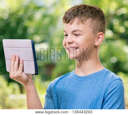 Outdoor portrait of happy teen boy 12-14 year old with tablet pc watching video in summer park