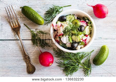 Healthy diet food: vegan vegetable salad with fresh cucumbers radish green onion black olives dill and feta cheese. In white ceramic bowl on light wooden background. Top view.