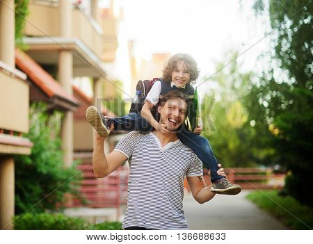 The father carries on his shoulders of his son-schooboy. Little learner with one arm hugs dad for the neck and the other holding a green folder. Father and son smiling happily. Back to school.