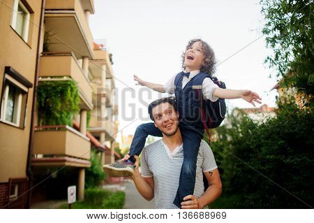 Little pupil sitting on the shoulders of his father. He placed his hands to the sides as if flying. Dad and son a great mood. They play on the way to school. Behind the schoolboy's backpack.