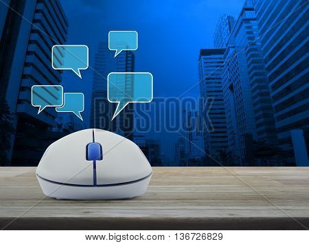 Wireless computer mouse with social chat sign and speech bubbles on wooden table in front of city tower background Social network concept