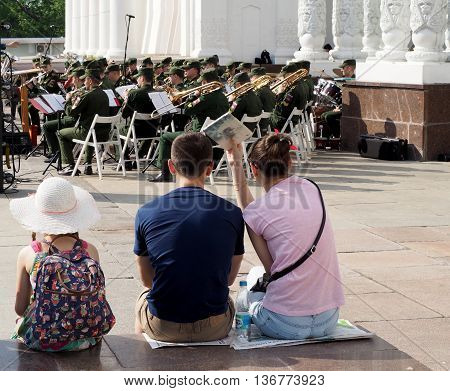 MOSCOW, RUSSIA - May 28, 2016: A pre-adolescent girl and a pair of young men seated on the parapet during a performance of the brass band. Central Avenue in the Park of VDNH Moscow. International Military Music Festival 