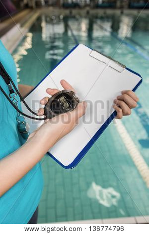 Mid section of coach with a stop watch and clipboard by pool at leisure center