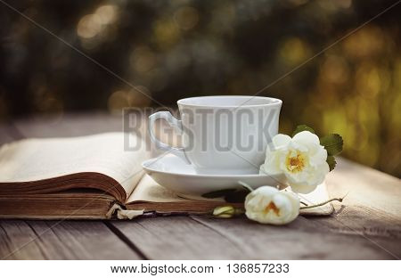 White cup with a flowers of wild rose on an open old book on a wooden table.