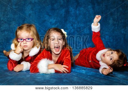 Three sisters lay on a portrait studio floor. One cooperates with the photographer and smiles one scratches at her eye and one makes silly faces as she rolls around the floor.