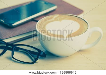 A cup of coffe with heart pattern in a white cup on smartphone leather notebook and eyeglasses on wooden table.Close up.Vintage tone