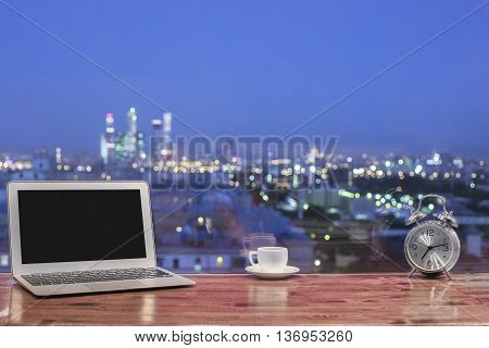 Front view of creative designer desktop with blank notebook silver alarm clock and coffee cup on blurry night city background. Mock up