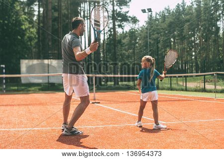 Training together. Full length rear view of father in sports clothing teaching his daughter to play tennis while both standing on tennis court