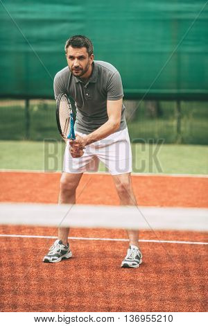 Practicing tennis. Confident young man in sports clothes carrying tennis racket and looking concentrated while standing on tennis court