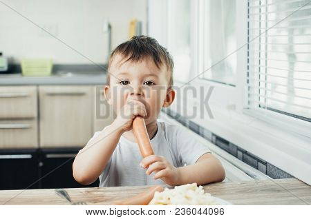 Handsome Boy Shaggy And Shkod Eats Sausage In The Kitchen In A White T-shirt In The Afternoon