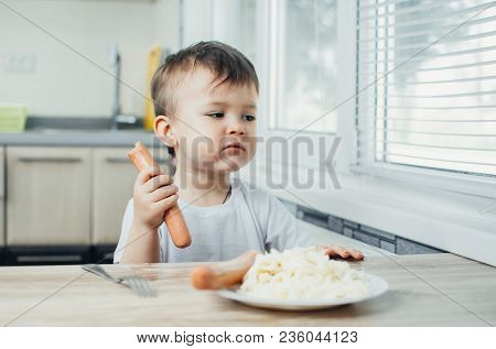 Handsome Boy Shaggy And Shkod Eats Sausage In The Kitchen In A White T-shirt In The Afternoon