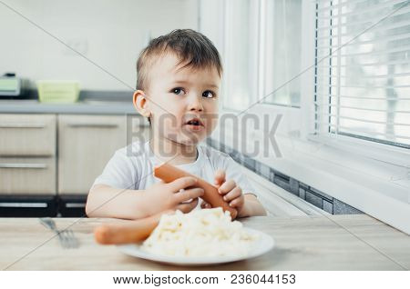 The Child Is Sitting In The Kitchen And Greedily Eats Sausage