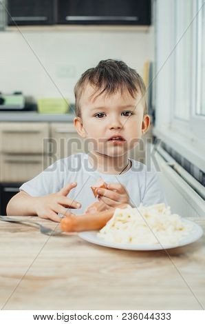 The Child Is Sitting In The Kitchen And Greedily Eats Sausage