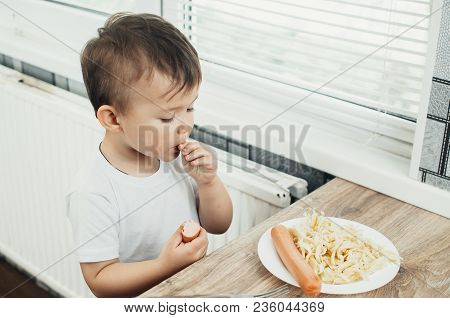 The Child Is Sitting In The Kitchen And Greedily Eats Sausage