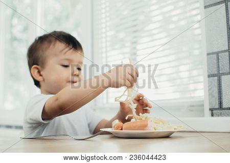 The Child Is Sitting In The Kitchen And Greedily Eats Sausage