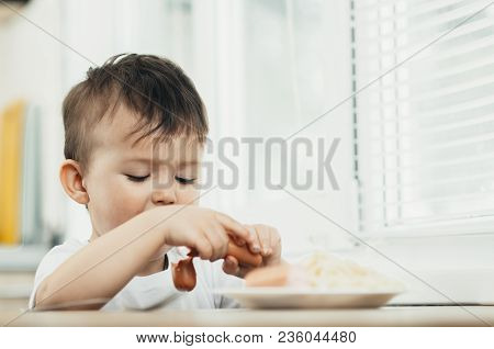The Child Is Sitting In The Kitchen And Greedily Eats Sausage