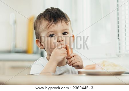 The Child Is Sitting In The Kitchen And Greedily Eats Sausage