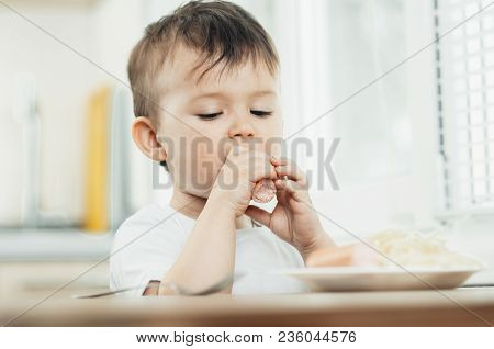 The Child Is Sitting In The Kitchen And Greedily Eats Sausage