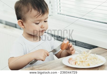 The Child Is Sitting In The Kitchen And Greedily Eats Sausage
