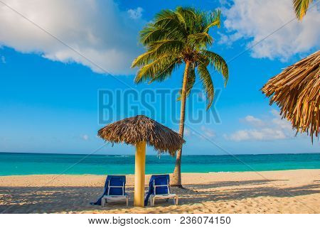 Holguin, Cuba, Playa Esmeralda. Umbrella And Two Lounge Chairs Around Palm Trees. Tropical Beach On 