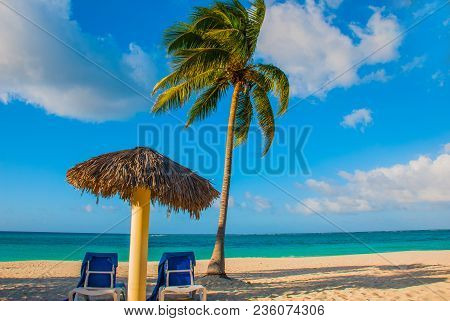 Holguin, Cuba, Playa Esmeralda. Umbrella And Two Lounge Chairs Around Palm Trees. Tropical Beach On 
