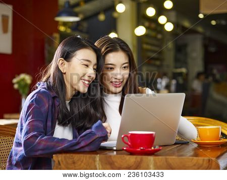 Two Happy Young Asian Women Sitting In Coffee Shop Looking At Laptop Computer Together.