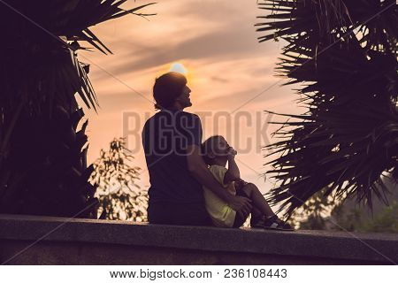 Silhouettes Of The Father And Son, Who Meet The Sunset In The Tropics Against The Backdrop Of Palm T