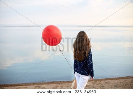 Portrait Of Young Woman With Red Air Balloon And Present Bag Near The Calm Sea Or Lake. Clouds Are R