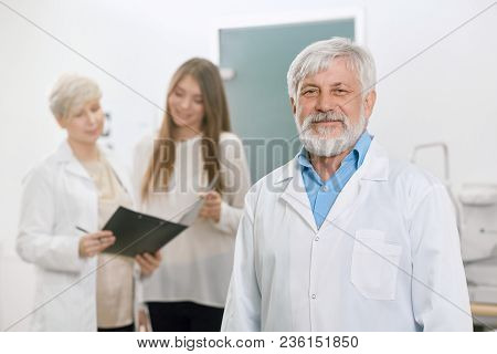 Confident Old Doctor Staying In Front Of Assistant And Patient. Looking Expirienced, Skilled, Profes