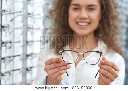 Smiling, Curly Oculist Wearing White Uniform Giving Glasses To Try On. Doctor Is Standing In Front O