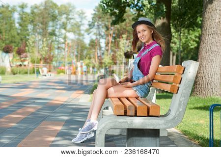 Young Pretty Girl Sitting On The Benchin The City Park Keeping A Book. Wearing Blue Jeans Shorts, Br