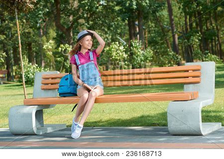 Young Stylish Dressed Girl Sitting On The Benchin The City Park Keeping A Book. Wearing Blue Jeans S