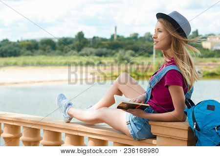 Pretty Young Student Sitting With A Book Near River In The City. Looking Relaxed, Happy And Satisgie