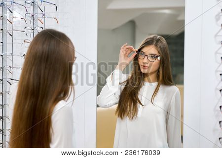 Young Girl Trying On Eyeglasses In Front Of Mirror. Helping To Improve Vision, Saving Eye Health. We