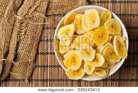 Heap Of Dried Banana Chips Snack In White Bowl On Bamboo Mat With Burlap On Table