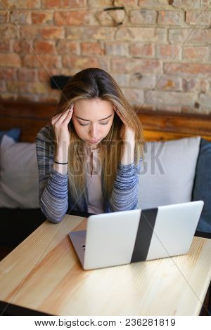 Young Woman Sitting In Cozy Coffee House Near Wooden Table With Glass Cup Of Cappuccino And Using La