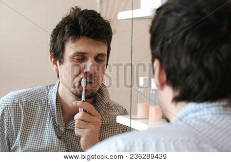 Tired Sleepy Man With A Hangover Who Has Just Woken Up Brush His Teeth, Looks At His Reflection In T